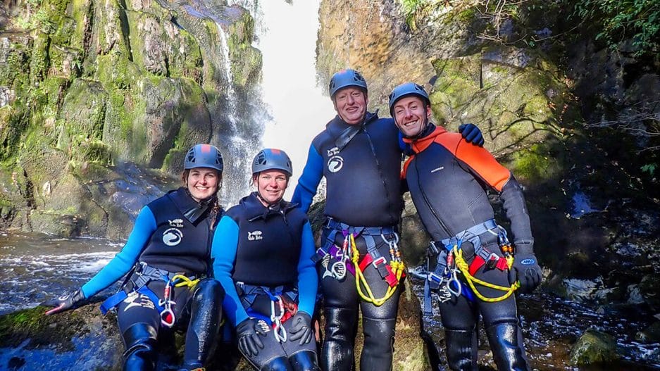 Four people dressed in wetsuits and canyoning equipment are posing in front of a waterfall, they all look happy canyoneers.